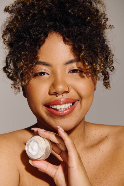 Belle femme afro-américaine aux cheveux bouclés tenant un pot de crème dans un décor de studio, se concentrant sur les soins de la peau. - Photo, image