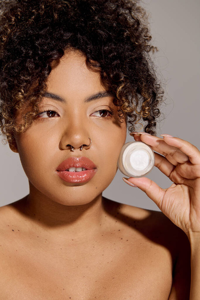 Uma jovem afro-americana com cabelo encaracolado segura um frasco de creme na frente de seu rosto, enfatizando cuidados com a pele e beleza. - Foto, Imagem