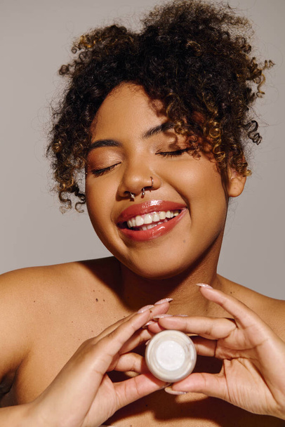 Belle femme afro-américaine tenant un pot de crème aux cheveux bouclés, mettant l'accent sur les soins de la peau et la routine de beauté. - Photo, image