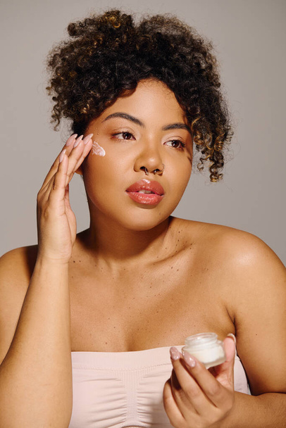 Una joven afroamericana con el pelo rizado sosteniendo un frasco de crema delante de su cara. - Foto, Imagen