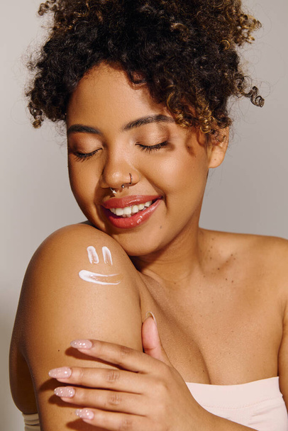 Güzel, kıvırcık saçlı, genç bir Afrikalı Amerikalı kadın stüdyo ortamında kolları ışıl ışıl parlarken gülümsüyor.. - Fotoğraf, Görsel