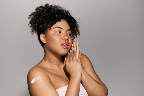 Eine junge Afroamerikanerin mit lockigem Haar in einem schulterfreien Oberteil hält sich in einem Studio zart die Hand vor das Gesicht. - Foto, Bild