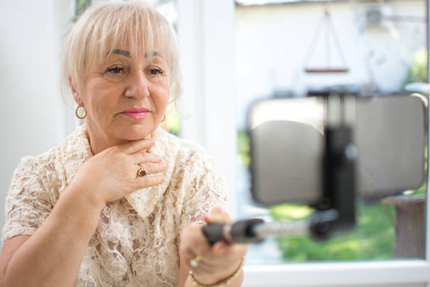 Mujer madura sosteniendo palo selfie con teléfono inteligente mientras tiene una videollamada en casa - Foto, Imagen