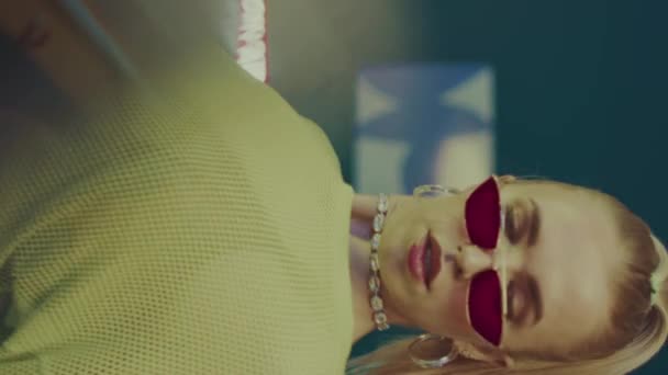 Verticale medio scatto di attraente giovane donna bionda in occhiali rosa vintage e y2k vestito danza seducente sulla macchina fotografica in camera fumosa - Filmati, video