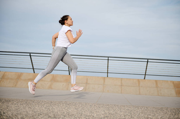 Retrato lateral de una mujer corriendo por el puente de la ciudad, haciendo footing matutino, haciendo ejercicio al aire libre. Atlética hembra quemando calorías y perdiendo peso mientras corre trotando corriendo en el paseo marítimo - Foto, imagen
