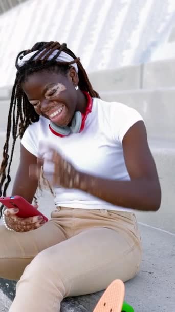 Joven adolescente africana con vitiligo divertirse utilizando el teléfono móvil sentado al aire libre. Concepto de cultura y tecnología juvenil. Vídeo HD vertical - Metraje, vídeo