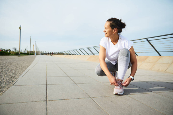 Mladá žena si váže tkaničky na růžových teniskách, usmívá se a připravuje se na ranní běhání venku ve městě. Běhání a běhání. Vědomí běžící maratonský koncept. Sport. Vytrvalost - Fotografie, Obrázek