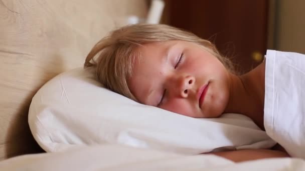 Mała dziewczynka z długimi włosami spokojnie śpi rano w wygodnym łóżku na białej poduszce. Zamknij klip slow motion. Idealny do projektów związanych z rodziną, dzieciństwem, lub - Materiał filmowy, wideo