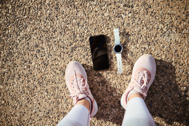 Pies de vista superior de una deportista en zapatillas deportivas de running rosa, un reloj de pulsera inteligente y un teléfono inteligente con pantalla digital de maqueta en blanco negro en el asfalto. Copiar el espacio publicitario para los deportes y la aplicación móvil fitness - Foto, Imagen