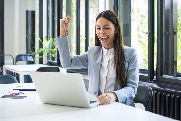 Happy excited woman working on laptop and triumphing with fist up in office. Achievement and satisfaction concept. - Photo, Image