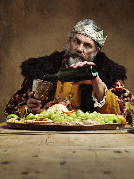 Koning, koninklijk en feest in tafel met wijn als heer op de eetzaal voor traditie, cultuur en maaltijd in het paleis. Portret, vorst en eten met man etend buffet of avondmaal met vertrouwen en kroon. - Foto, afbeelding