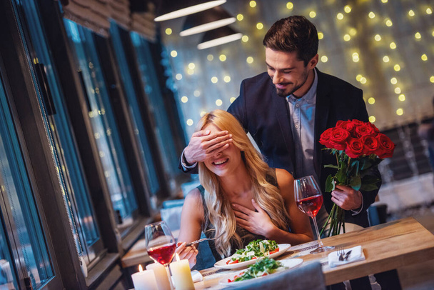 ハンサムなエレガントな男性がバラを保持しているし、レストランで驚きを作っている間彼のガール フレンドの目を覆っている、両方が笑っています。 - 写真・画像