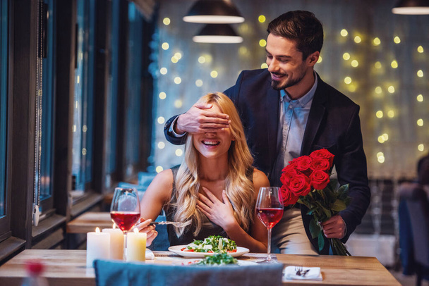 ハンサムなエレガントな男性がバラを保持しているし、レストランで驚きを作っている間彼のガール フレンドの目を覆っている、両方が笑っています。 - 写真・画像