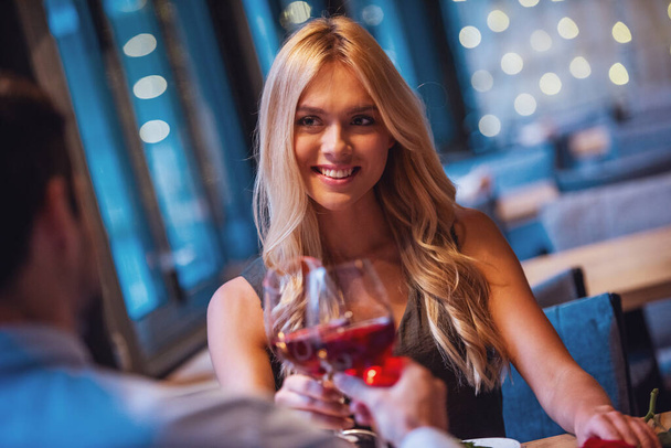 Όμορφη νεαρή γυναίκα βλέπει τον φίλο της, πίνοντας κρασί και χαμογελώντας ενώ έχοντας μια ημερομηνία στο εστιατόριο - Φωτογραφία, εικόνα