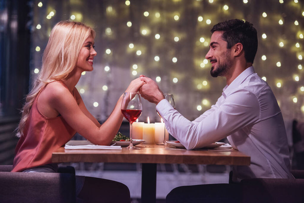 Piękna Młoda para jest patrząc na siebie, trzymając się za ręce i uśmiechając się podczas ich daty w restauracji - Zdjęcie, obraz