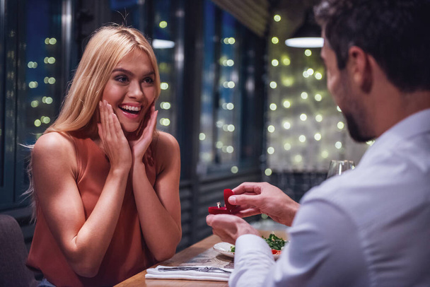 Красивая молодая женщина улыбается, пока ее парень держит обручальное кольцо и делает ей предложение в ресторане
 - Фото, изображение