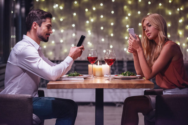 Νεαρό ζευγάρι χρησιμοποιώντας το smartphone τους κατά την ημερομηνία σε ένα εστιατόριο - Φωτογραφία, εικόνα