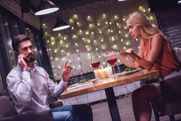 Όμορφη νεαρή γυναίκα είναι ενοχλημένος, ενώ ο φίλος είναι μιλάμε στο κινητό τηλέφωνο κατά την ημερομηνία σε ένα εστιατόριο - Φωτογραφία, εικόνα