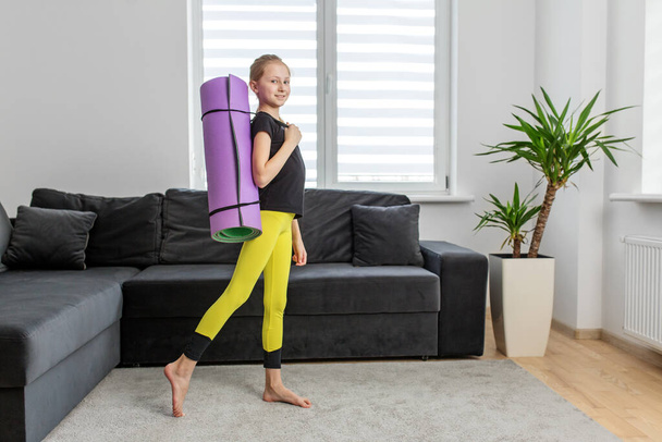 Jeune fille avec des promenades en queue de cheval avec tapis de yoga violet dans l'environnement à la maison, se préparant à la routine de remise en forme. - Photo, image