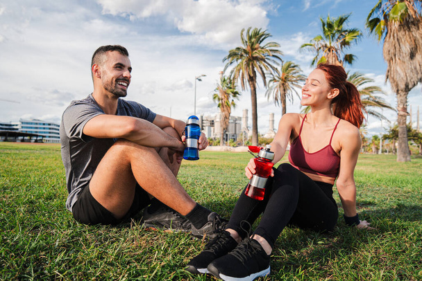 Sport couple talking and resting on the grass drinking wather after cardio workout. Tired young people recovering energy and having a conversation. Coach man and athletic woman refreshing together - Photo, Image