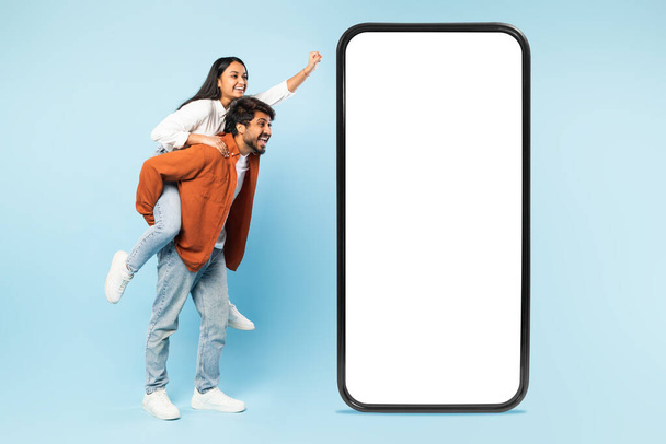 Mobile fun. Happy indian couple having fun near big cellphone with white blank screen for mockup, man giving piggyback ride to woman, advertising offer together, over blue background, collage - Photo, Image