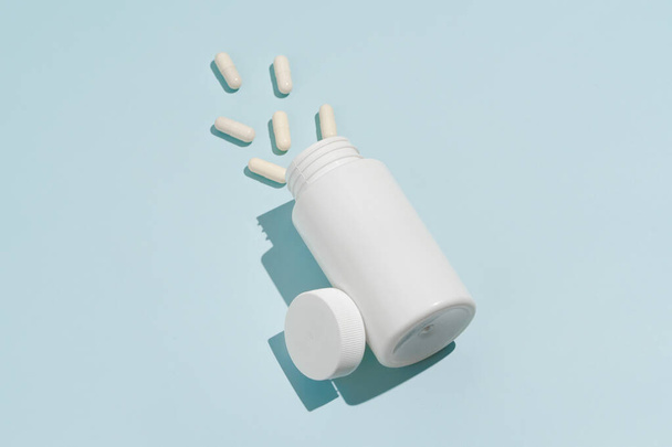 Белая банка-макет и пролитые таблетки капсулы наклонены на синий изолированный фон. Понятие фармации, медицины, лекарств от болезней, антибиотиков, здравоохранения. фон для вашего дизайна - Фото, изображение