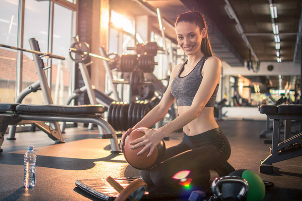 Спортивна дівчина тримає фітнес-кулю і розслабляється, сидячи на тренувальному килимку під час тренувальної перерви в тренажерному залі. - Фото, зображення