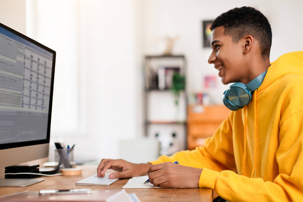 Vidám tinédzser srác fényes sárga kapucnis pulcsiban és fejhallgatóval mosolyog, miközben számítógépet használ, megragadja az élvezetes tanulás pillanatát otthon - Fotó, kép