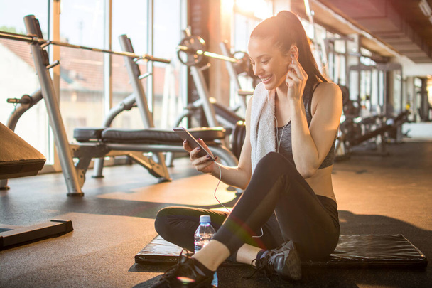 Beautiful young sportswoman with earphones listening to music on smart phone after sports training in gym. - Photo, Image