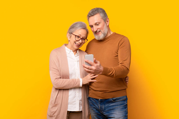 Onnellinen kypsä harmaa pari tarkastellaan älypuhelimen näytön, vanhempi mies ja nainen halaamalla käyttäessään mobiilisovellusta matkapuhelimeen, ostoksia tai websurfing verkossa, keltaisella taustalla - Valokuva, kuva