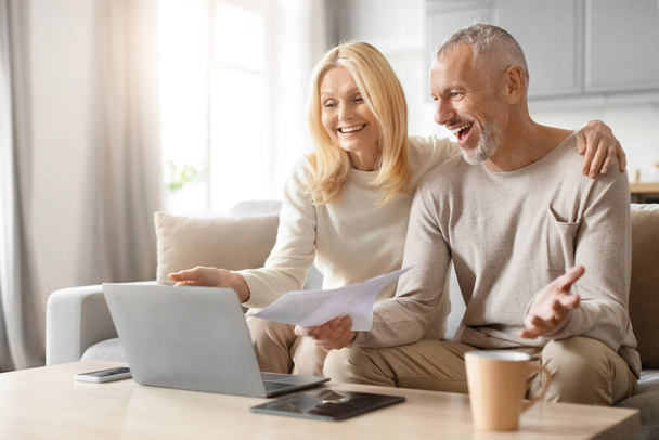 Lächelndes älteres Paar diskutiert glücklich mit Papieren und Laptop, sitzt auf einem Sofa in einem hellen Raum - Foto, Bild