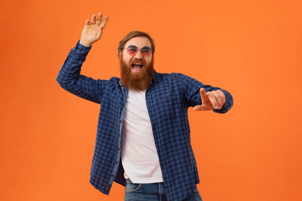 Счастливый рыжеволосый бородатый парень в повседневной одежде и солнечных очках, указывающих на тебя на фоне оранжевой студии. Европейский хипстер наслаждается вечеринками, танцами и весельем - Фото, изображение