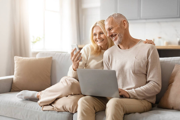 Старшая пара сидит на диване, смеясь, глядя на экран ноутбука, в хорошо освещенной гостиной - Фото, изображение