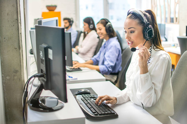 Beautiful female call center operator working on computer in office. Young customer support talking to customer with headset in the office. - Photo, Image