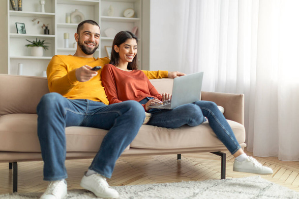 Un couple joyeux profite du temps libre sur un canapé, l'un avec un ordinateur portable et l'autre tenant une télécommande - Photo, image