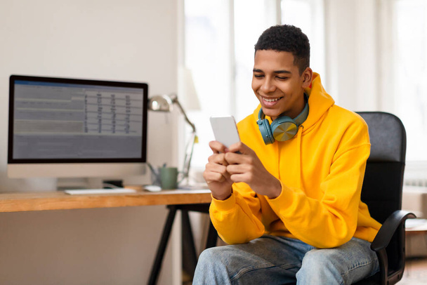 Un giovane allegro con una felpa gialla usa il suo smartphone con uno schermo di computer sullo sfondo - Foto, immagini