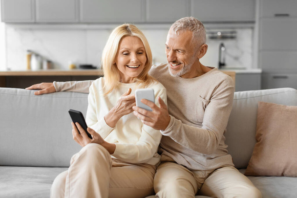 Веселая пожилая пара сидит на диване с комфортом, используя мобильный телефон, возможно, разделяя цифровой момент - Фото, изображение