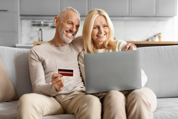 Зрелая пара счастливо использует кредитную карту, чтобы ходить по магазинам онлайн, сидя на диване дома - Фото, изображение