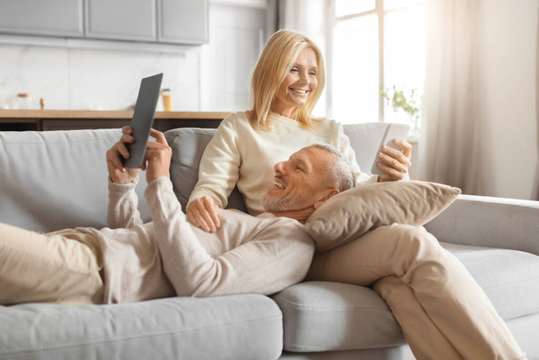 Старший чоловік сміється під час перегляду планшета, коли його партнер насолоджується своїм напоєм, обидва розділяють веселий момент - Фото, зображення