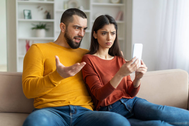 Een lastig echtpaar richt zich op een smartphone, hun uitdrukkingen suggereren bezorgdheid of schok - Foto, afbeelding