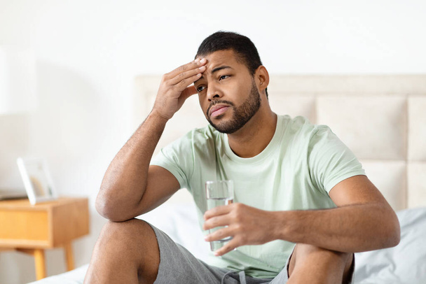 Hombre afroamericano sentado en la cama con la mano en la frente, sosteniendo un vaso de agua, expresando un dolor de cabeza o preocupación en un ambiente hogareño - Foto, imagen