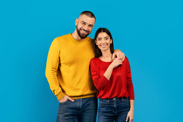 青い背景に自信を持って立っているカラフルなセーターを着た笑顔のカップル, 明るい背景に対して一緒にスタジオでポーズロマンと女性, カメラを見て, コピースペース - 写真・画像