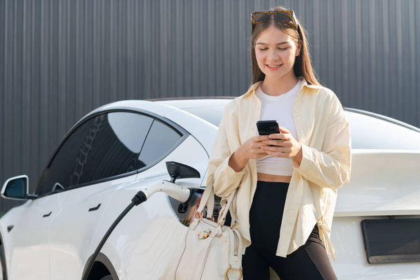 Nuori nainen tilalla ostoskassi ja käyttää älypuhelinta maksaa sähkön lataamiseen EV auton akku latausasemalta kaupungin kauppakeskuksen parkkipaikalla. Moderni nainen käy ostoksilla ekoautolla. Kuuluisa - Valokuva, kuva
