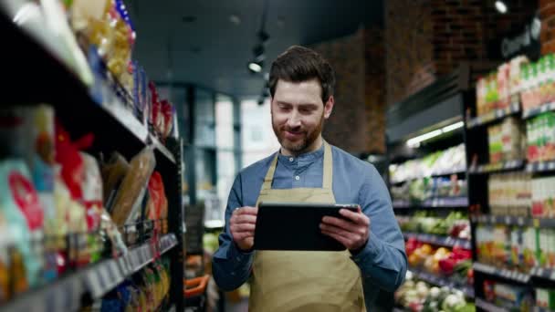 Portrait d'un homme souriant debout parmi les allées des supermarchés et défilant à travers un gadget moderne avec stylet numérique. Vendeur senior vérifiant la qualité du travail des employés avec satisfaction. - Séquence, vidéo