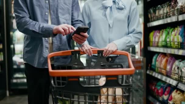 Twee mensen in de supermarkt. Blanke man met smartphone terwijl jonge vrouw in de buurt komt en een winkelwagen vol met enkele artikelen duwt. Concept van aankoop, technologie en winkelen. - Video