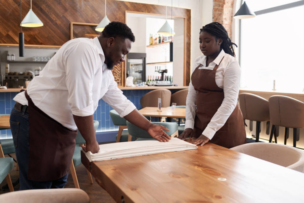 Junge afroamerikanische Oberkellnerin bringt Kellnerin bei, wie man Tischdecken bei der Arbeit in modernen - Foto, Bild