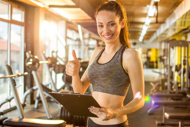 Cheerful female personal trainer with clipboard showing thumb up at gym. - Photo, Image