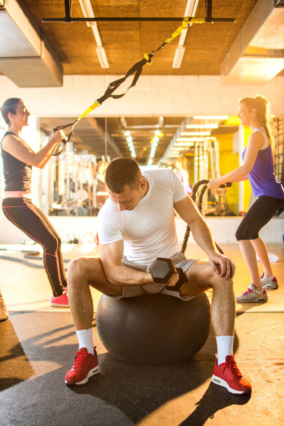 Hübscher junger Mann hebt Gewicht, während er auf einem Fitnessball sitzt und im Hintergrund Menschen trainiert. - Foto, Bild