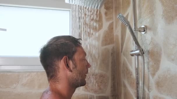 浴室でリフレッシュするために雨シャワーの下で涼しい水で洗面頭と体を取る男. 朝のルーチンとボディケアの衛生。 ハンサムな日焼けした男がシャワーを浴びた. サイドビュー - 映像、動画