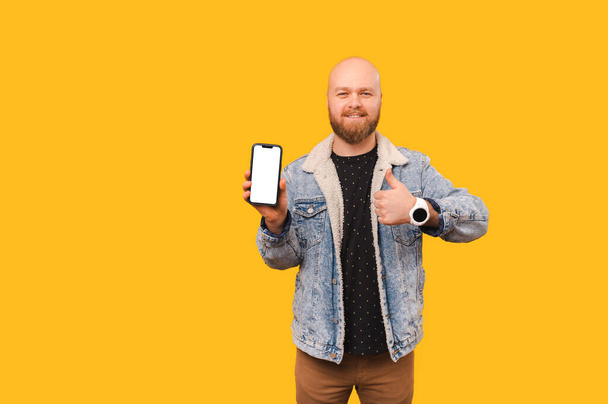 Beau chauve barbu homme tient un téléphone avec écran blanc et pouce vers le haut sur fond jaune. - Photo, image